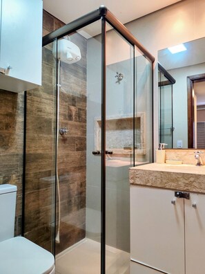 A modern bathroom with a glass shower enclosure, a toilet, and a vanity with a marble countertop. - Condomínio Praias do Guarujá - Praia do Tombo - SP (Guarujá)