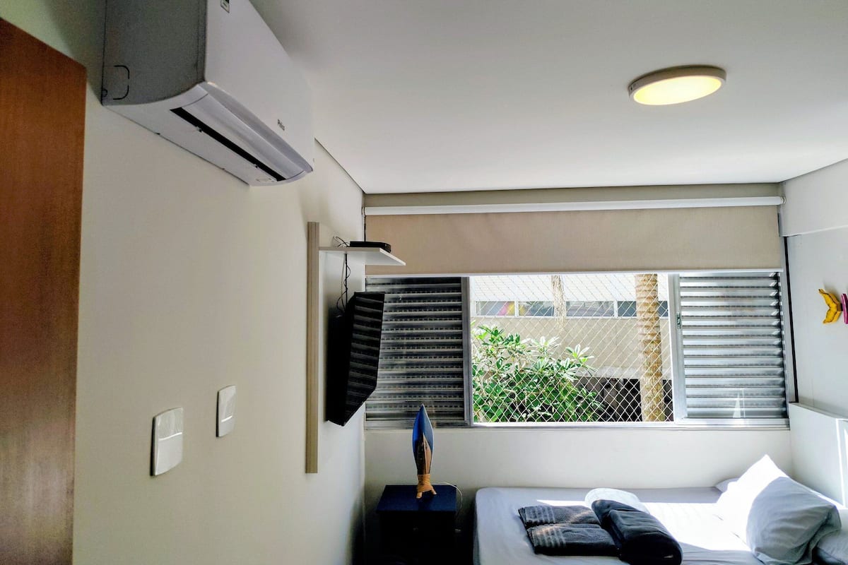 Ein Hotelzimmer mit Bett, Nachttisch, Klimaanlage, einem Fenster mit Jalousien und Blick auf Grünfläche.