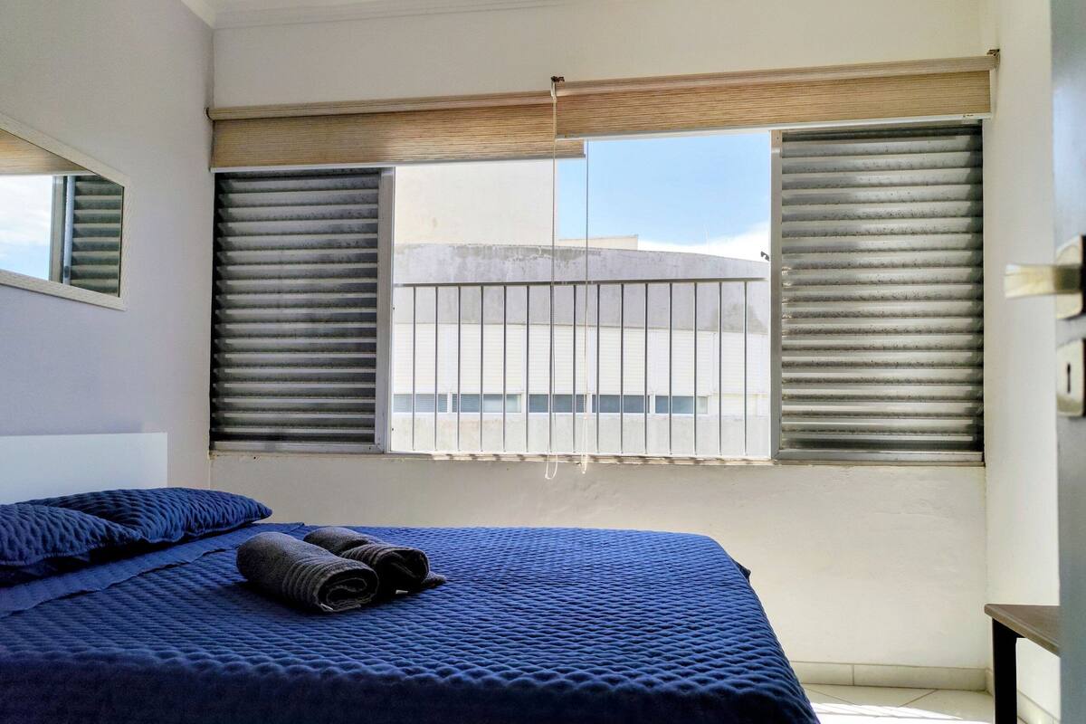 Ein Schlafzimmer mit einem Bett, zwei Fenstern mit Jalousien, Blick auf ein Gebäude und einem kleinen Tisch.