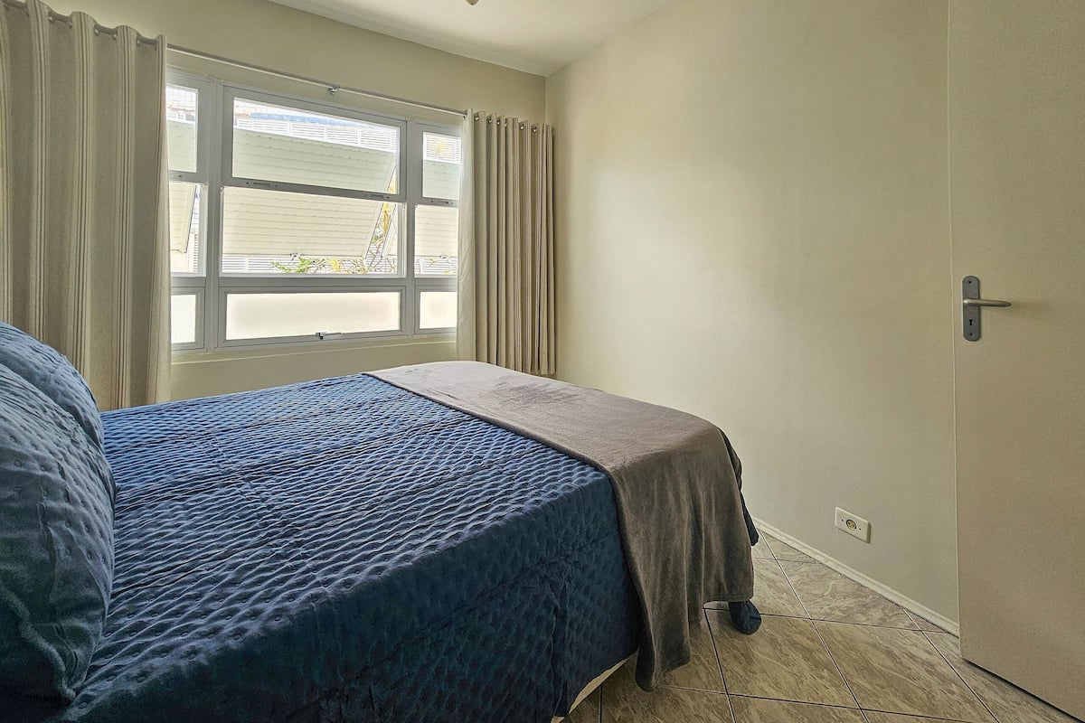 Ein Schlafzimmer mit Bett, Fenster mit Vorhängen, einer Tür und einem Fliesenboden.