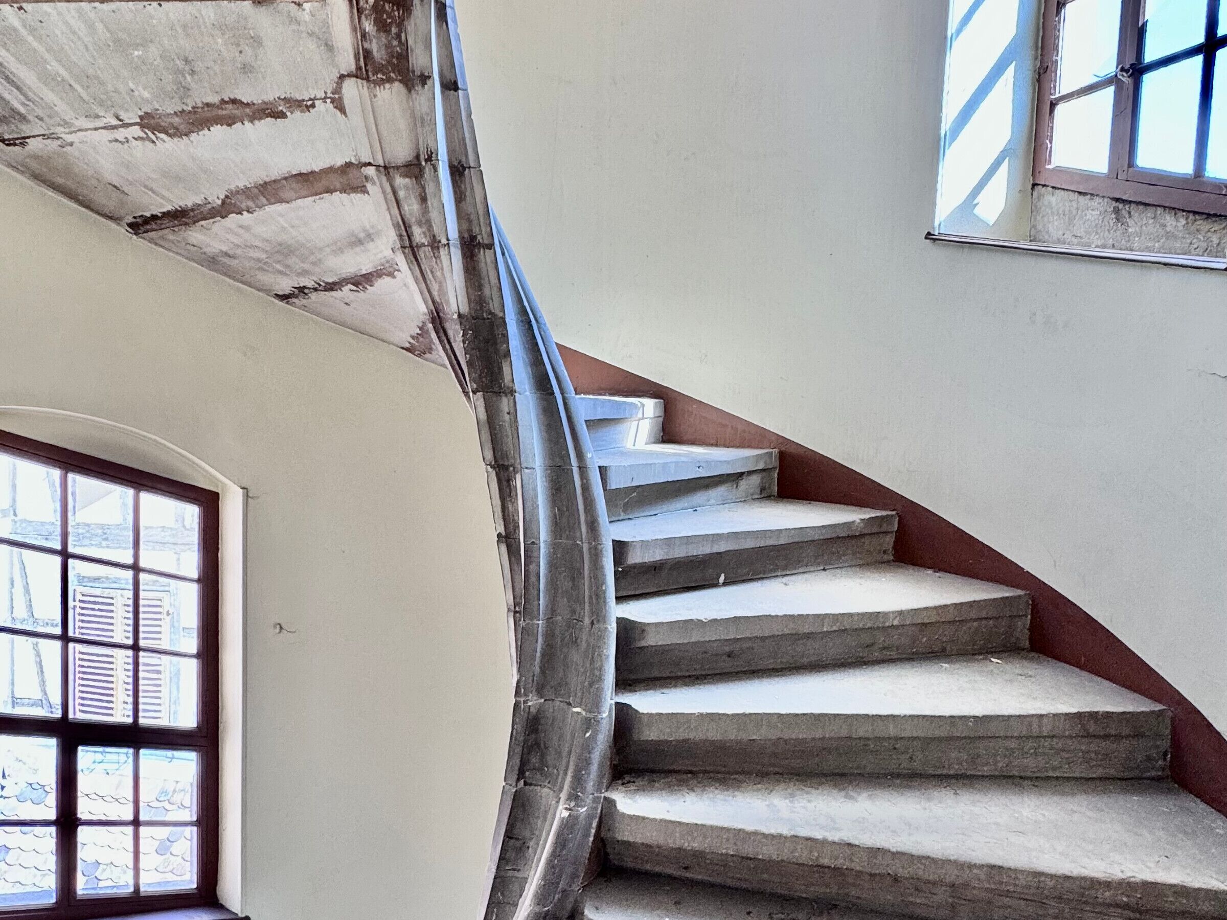 A spiral staircase with concrete steps and a metal handrail, leading to a landing with a window.