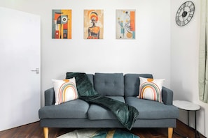 A grey sofa with two pillows, a green throw blanket, and three abstract paintings on the wall. - Charming 1-BR Apartment near Brick Lane Sleeps 3 (Greater London)