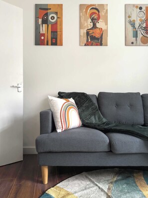 A grey sofa with a colorful pillow, a dark throw blanket, and a patterned rug in a living room with abstract paintings on the wall. - Charming 1-BR Apartment near Brick Lane Sleeps 3 (Greater London)
