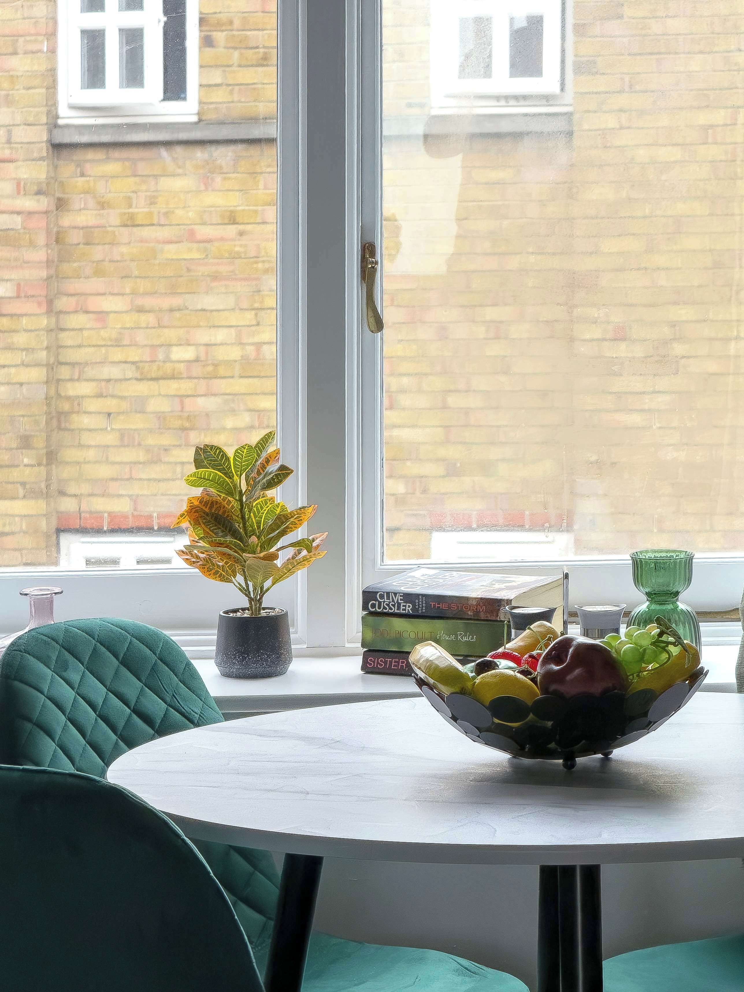A room with a round table, a potted plant, a stack of books, and a fruit bowl.