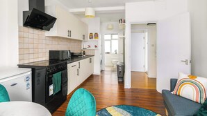 A modern kitchen with a black stove, microwave, and a white refrigerator. There is a dining area with teal chairs and a colorful rug. - Charming 1-BR Apartment near Brick Lane Sleeps 3 (Greater London)