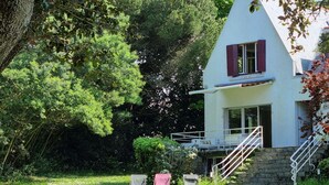 Ein weißes Gebäude mit roten Fensterläden, einer Steintreppe und einem Rasenstück mit zwei Stühlen.
