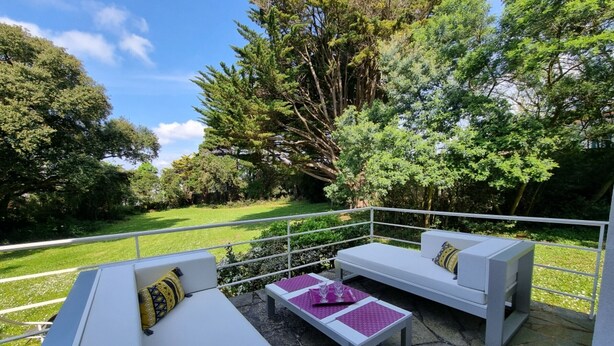 Eine Terrasse mit weißer Gartenmöbelgruppe, darunter Sofas und ein Tisch, auf einem Steinboden mit Blick auf eine grüne Landschaft und Bäume.