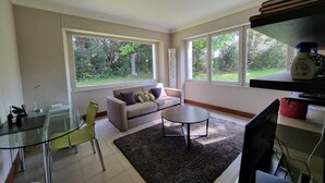 Ein modernes Wohnzimmer mit einem Glastisch, einem grauen Sofa und einem großen Fenster mit Blick ins Grüne.
