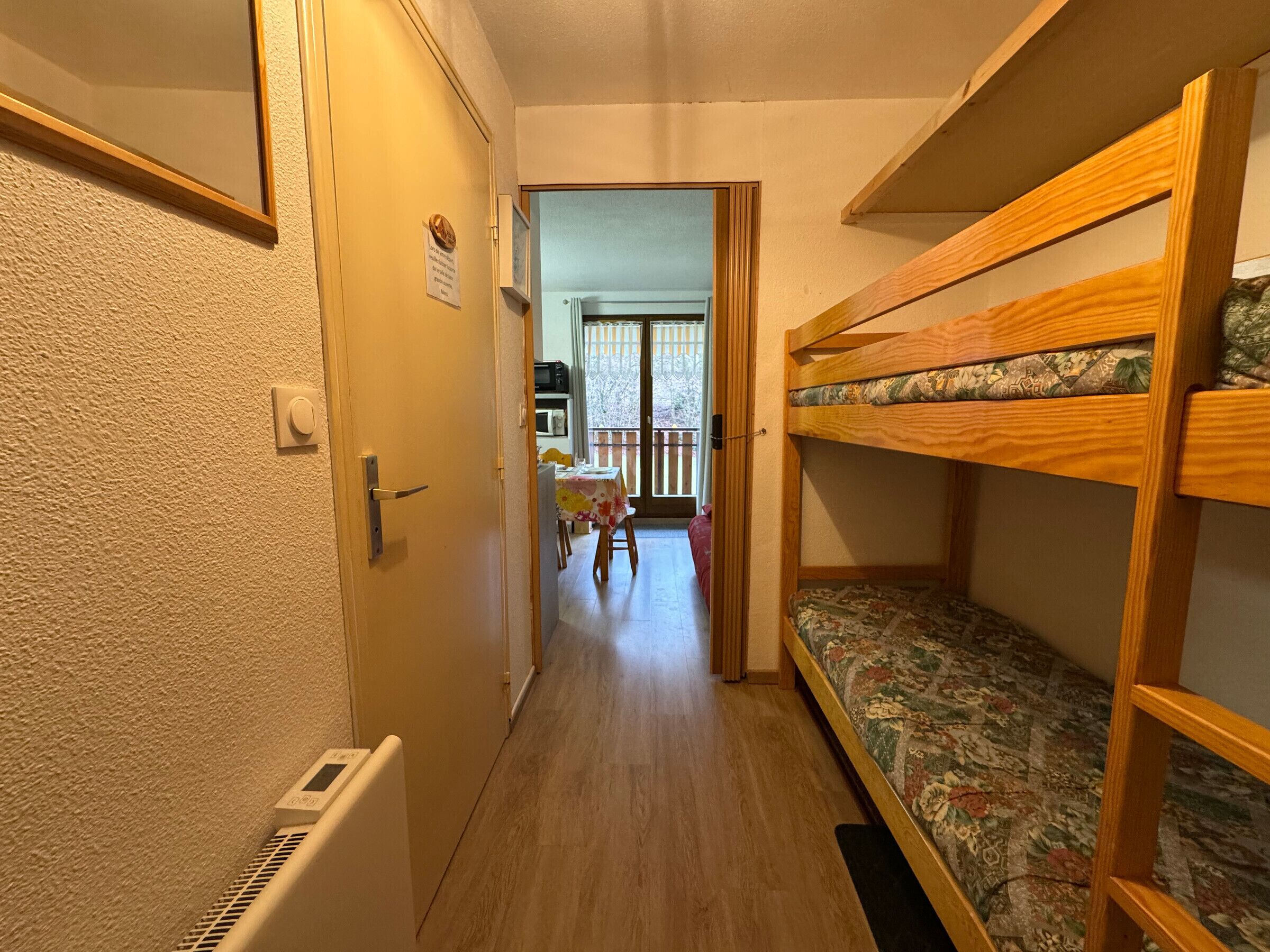 A narrow hallway with a wooden bunk bed on the right, a door on the left, and a table with chairs in the background.