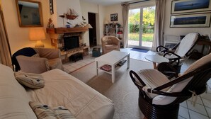 Ein Wohnzimmer mit Kamin, einer Couch, einem Couchtisch und einem Sessel. Ein Bücherregal und ein Fenster mit Vorhängen sind ebenfalls vorhanden.