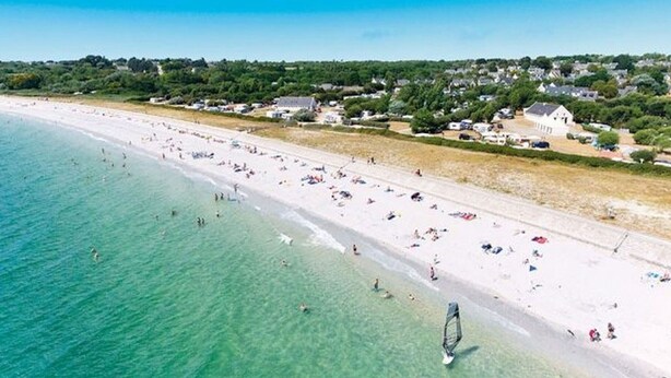 En strand med kristallklart turkost vatten, vit sand och några byggnader i bakgrunden.