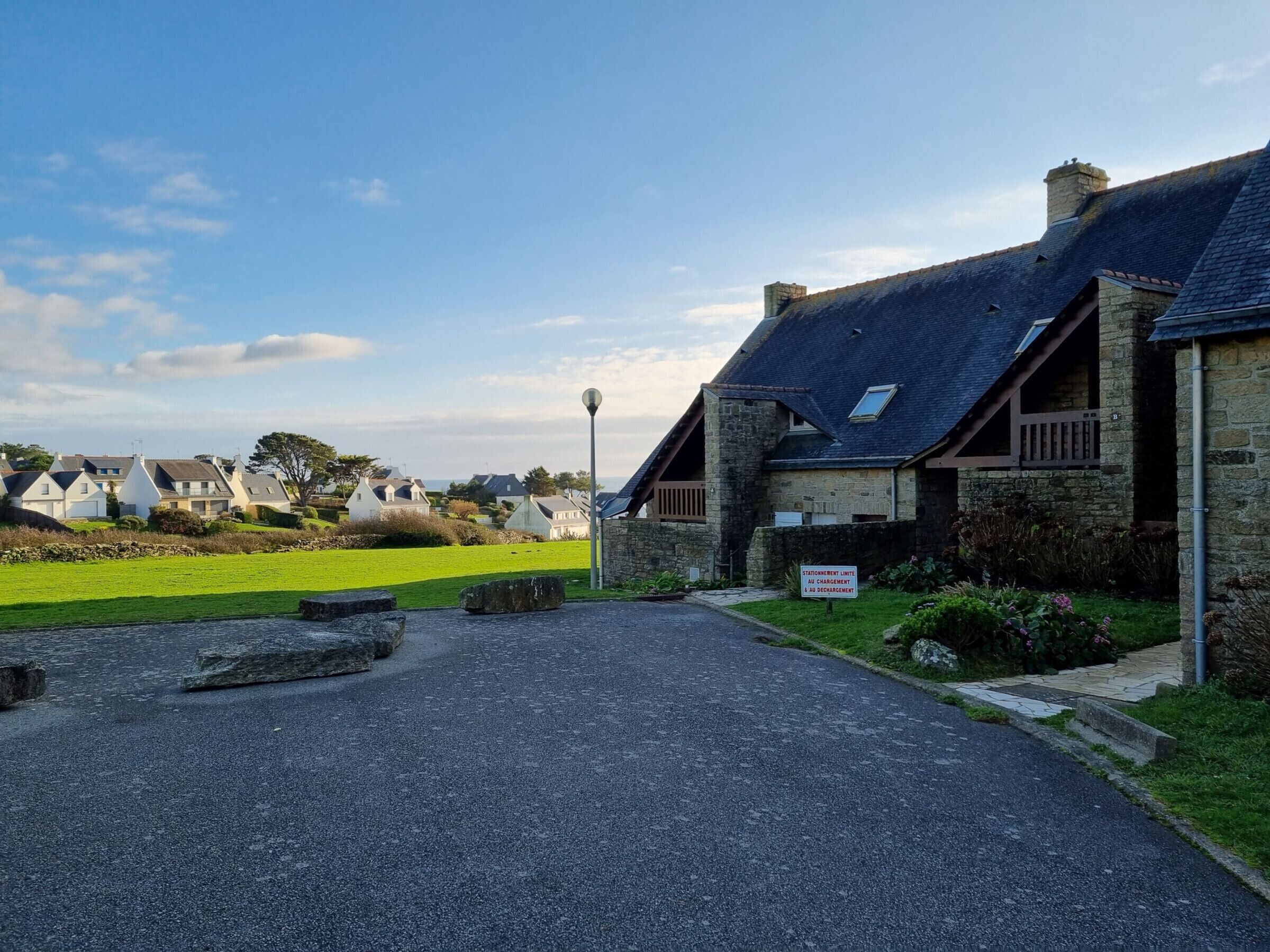 A stone building with a sloped roof, a paved driveway, and a grassy area with a sign indicating a parking area.