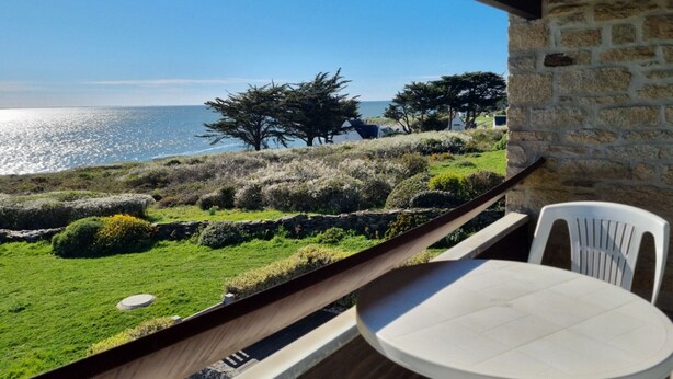 Un balcone con tavolo e sedie che si affaccia su un paesaggio costiero con alberi e vista sul mare.