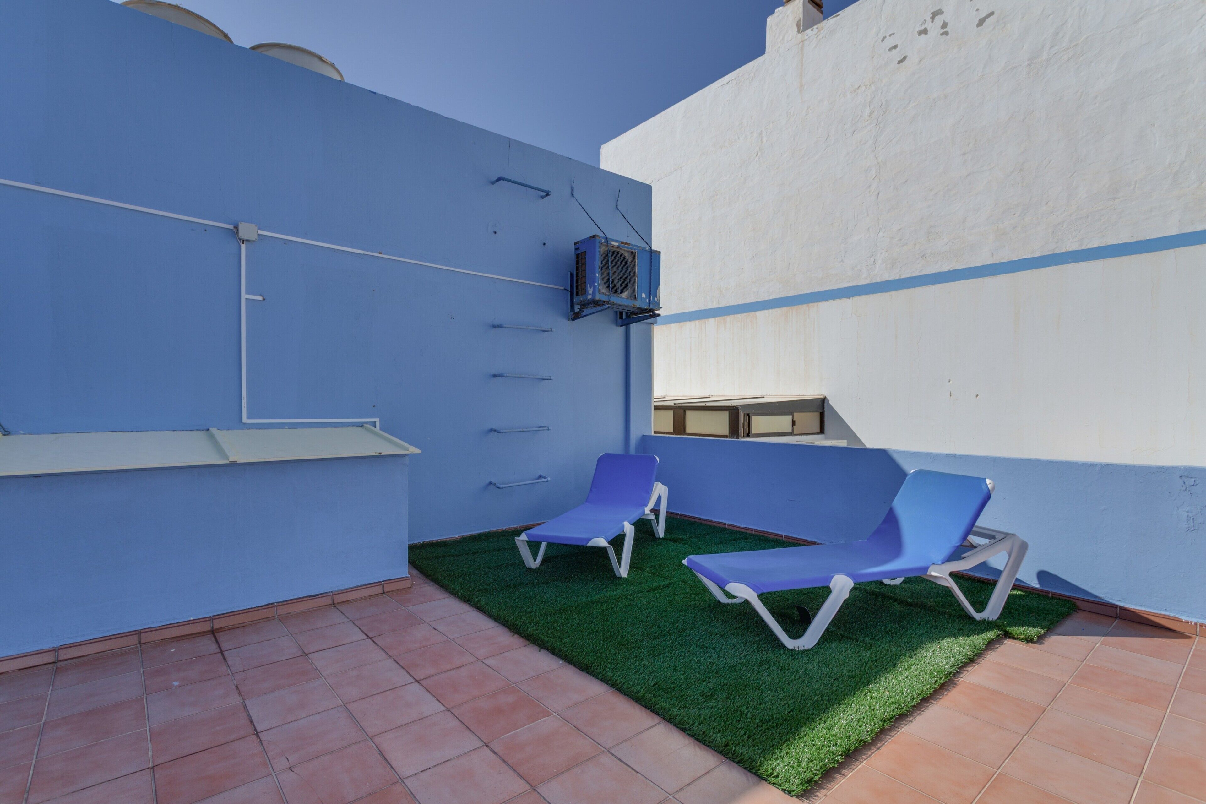 Pared exterior azul y blanca con un equipo de aire acondicionado pequeño, dos sillones azules y una zona de césped artificial verde.