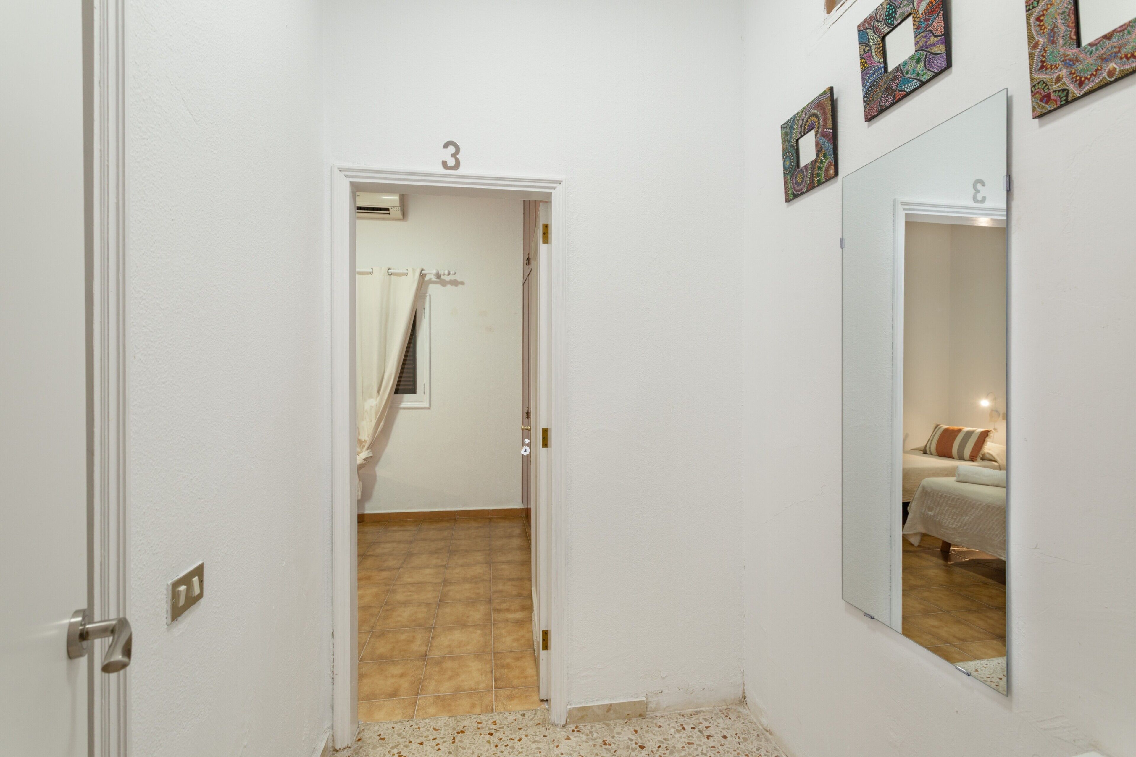Un pasillo con una puerta marcada '3', un espejo que refleja una cama y un suelo con baldosas.