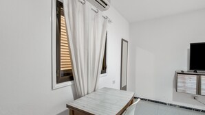 Une pièce comprenant une table, une fenêtre avec des stores, un appareil de climatisation, un miroir et un téléviseur.