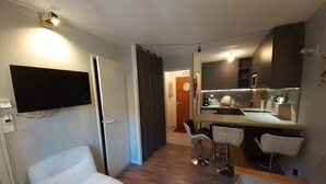 A compact living space with a flat-screen TV mounted on the wall, a kitchenette with a sink and countertop, and a seating area with two white chairs and a sofa. - Studio with Cabin in Valfréjus - Pets Allowed (Valfréjus)
