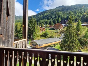 Varanda de madeira com detalhes em formato de coração, com vista para uma área montanhosa com edifícios e árvores.