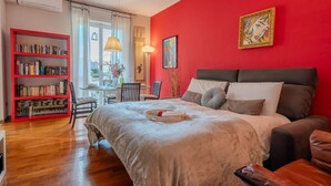 A bedroom with a bed, a red wall, a bookshelf, and a window.