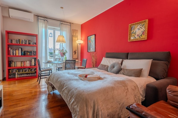 A bedroom with a bed, a red wall, a bookshelf, and a window.