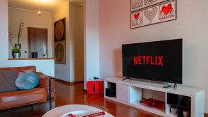 A living room with a leather sofa, a white coffee table, a flat-screen TV displaying 'NETFLIX,' and a wall with framed artwork.