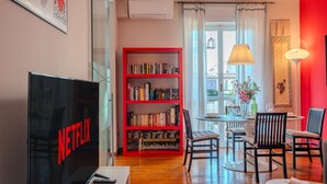 A living room with a bookshelf, a dining table, chairs, a TV, and a window with curtains.