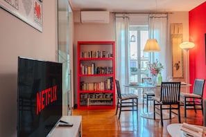 A living room with a bookshelf, a dining table, chairs, a TV, and a window with curtains.