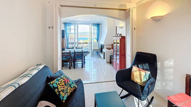 Uma sala de estar com sofá, poltrona e mesa. Há uma área de refeições com mesa e cadeiras ao fundo.