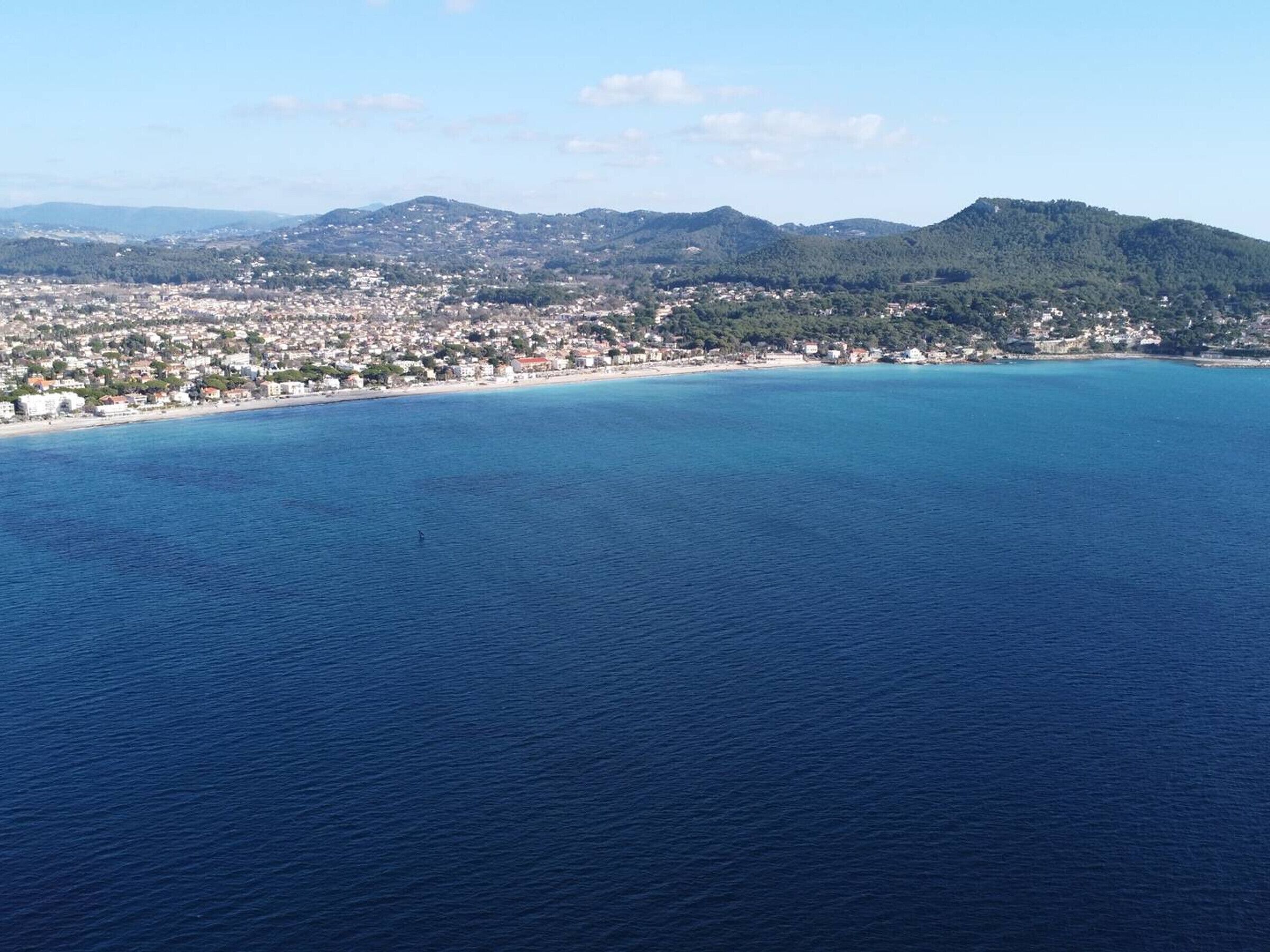 Vista aérea de uma cidade costeira com praia, casas e montanhas ao fundo.