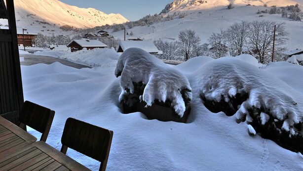 Paesaggio montano innevato con una baita di legno e un tavolo con sedie in primo piano.