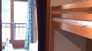Et værelse med en køjeseng i træ, et lille bord med en lampe, og udsigt til en balkon med mønstrede gardiner.