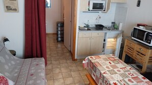 Et kompakt køkken med mikroovn, vask og skabe. Et spisebord med et mønstret duge er i forgrunden.