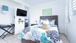 A modern bedroom with a bed, a flat-screen TV, a small shelf with a plant, and a painting on the wall. - Room-mar Azul - Ocean View Private Studi (Arecibo)