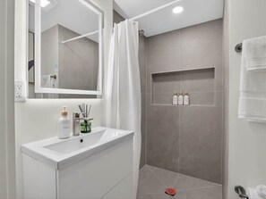 Un baño moderno con tocador blanco, un espejo grande, zona de ducha con puerta de cristal y repisa con artículos de aseo.