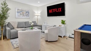 Ein modernes Wohnzimmer mit einem grauen Sofa, weißen Sesseln, einem Couchtisch, einem Billardtisch, einem Fernseher mit dem Schriftzug 'NETFLIX' und einer Karte von 'BOSTON' an der Wand.