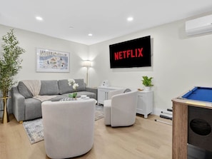 Una sala de estar moderna con un sofá gris, sillones blancos, una mesa de centro, una mesa de billar, un televisor mostrando 'NETFLIX' y un mapa de 'BOSTON' en la pared.