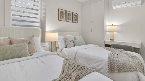 Ein Hotelzimmer mit zwei Betten, einem Fenster mit Jalousien, einem Nachttisch mit Lampe und einer Klimaanlage.