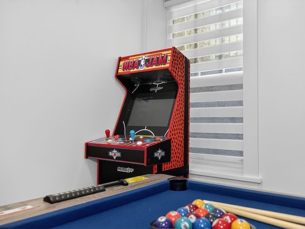Una mesa de billar con una máquina recreativa roja y negra con el rótulo 'NBA JAM' en una habitación con paredes blancas y una ventana con persianas.