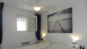 Un dormitorio con cama, ventilador de techo, ventana con cortinas, mesita de noche con lámpara y un cuadro colgado en la pared.