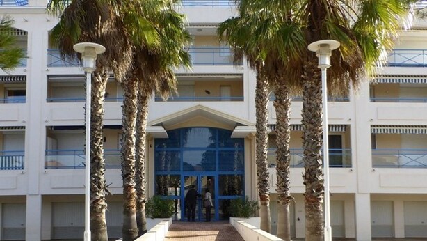 Un edificio con entrada central, palmeras y un camino pavimentado.