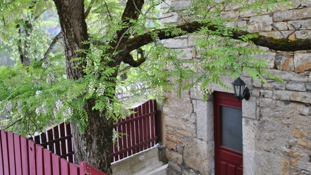 A stone building with a red door, a small table with a patterned tablecloth, and a red railing. - Gîte “Le Vignou” in Ste-Enimie: Mountain View, Private Terrace, and Wi-Fi (Gorges du Tarn Causses)