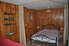 Ett sovrum med träpanel på väggarna, en sängram i metall, ett nattduksbord med en lampa och en hylla med böcker.