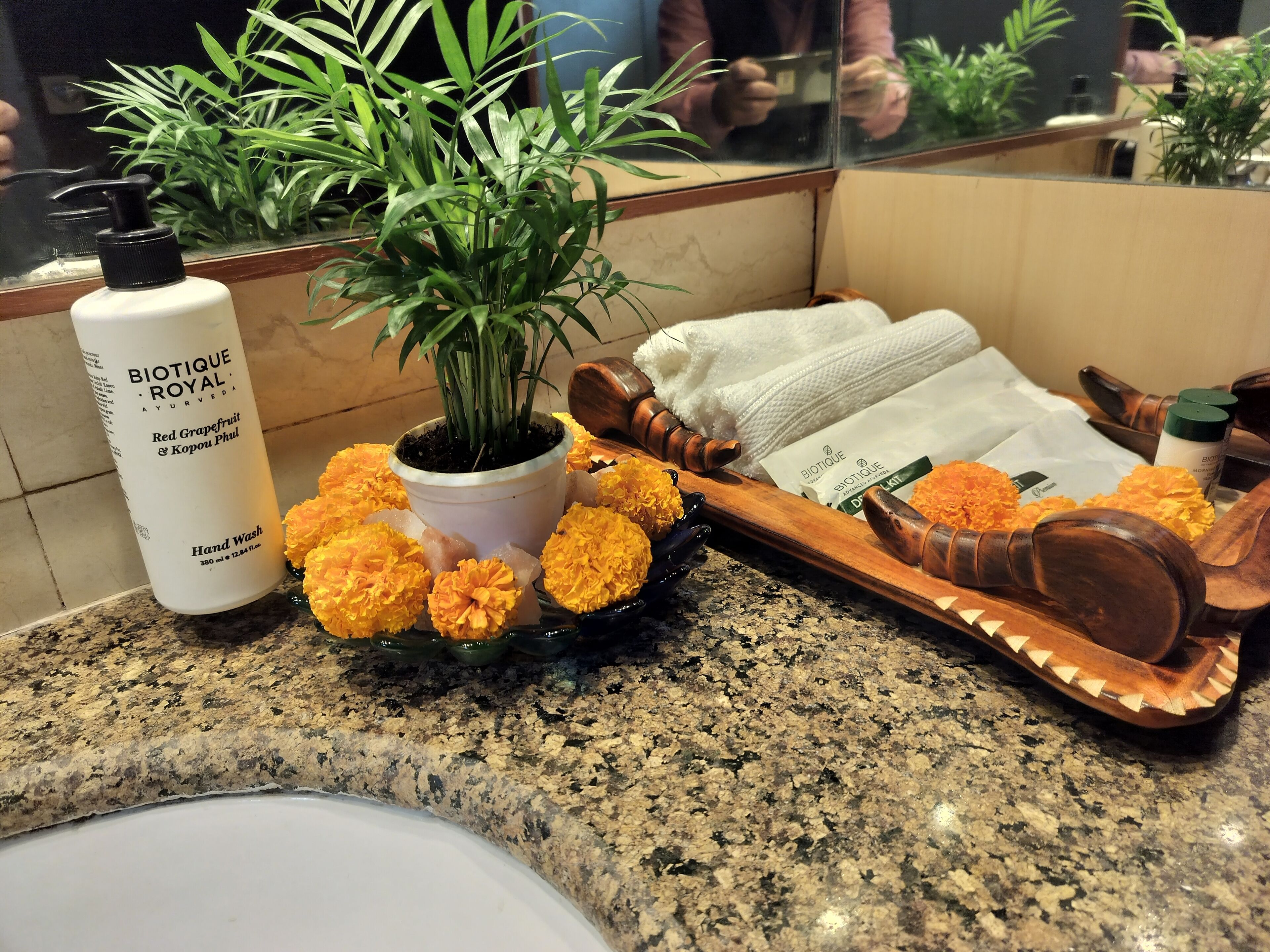 A bathroom counter with a hand wash bottle, a potted plant, a tray with flowers, and a towel.