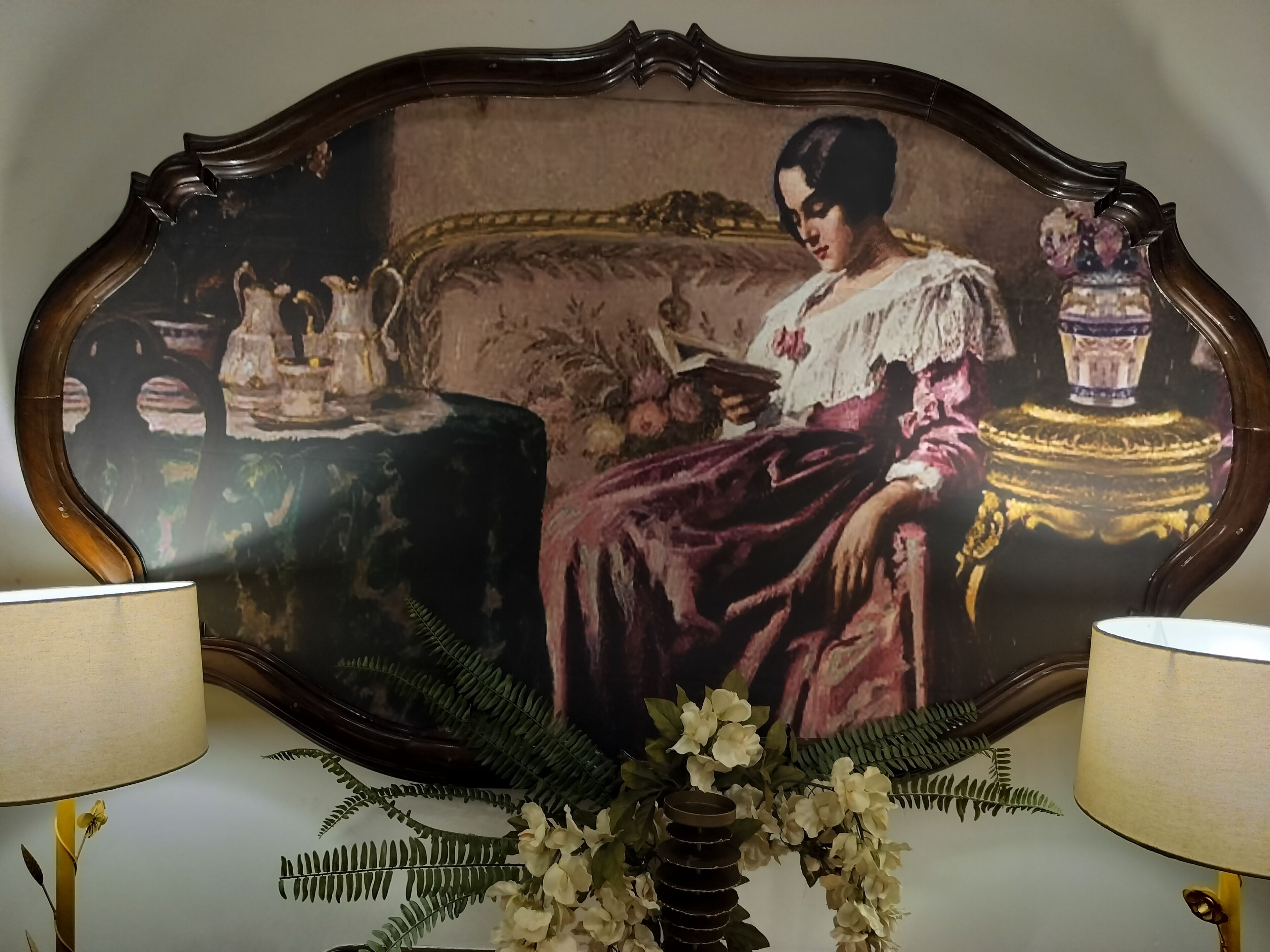 Un cuadro enmarco que muestra a una mujer con vestido rosa sentada en un sofá con un libro, rodeada de objetos decorativos.