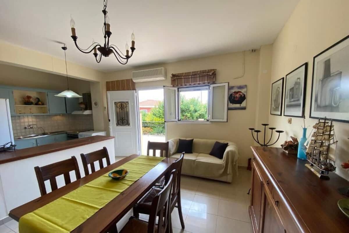 Un coin repas avec une table et des chaises en bois, un lustre, un canapé et, au fond, une cuisine.