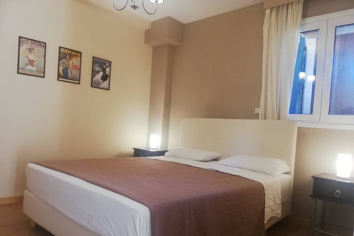 Une chambre à coucher avec un lit, une table de chevet, une fenêtre avec des rideaux et deux affiches au mur.