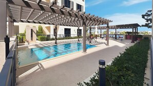 A swimming pool with a pergola and lounge chairs, surrounded by a landscaped area and a building with balconies. - Stylish 2BR with Beach Access (Dubai)
