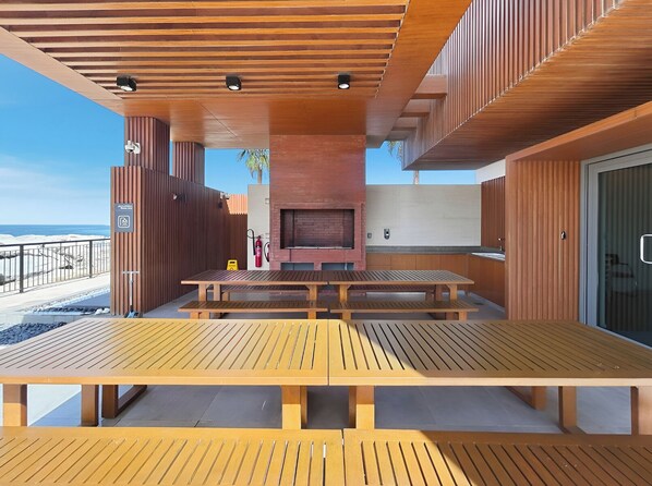 An outdoor patio with wooden tables and benches, a brick fireplace, and a view of the beach. - Stylish 2BR with Beach Access (Dubai)