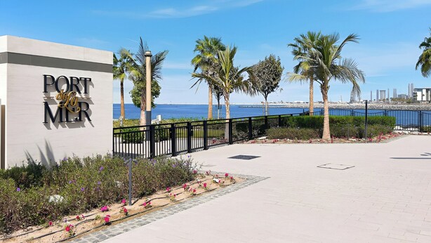 A coastal walkway with palm trees, a sign reading "Port de la Mer", and a view of the sea and city skyline. - Stylish 2BR with Beach Access (Dubai)