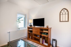 Une pièce avec un téléviseur posé sur un meuble en bois, une fenêtre avec une plante, un radiateur et une table en verre.